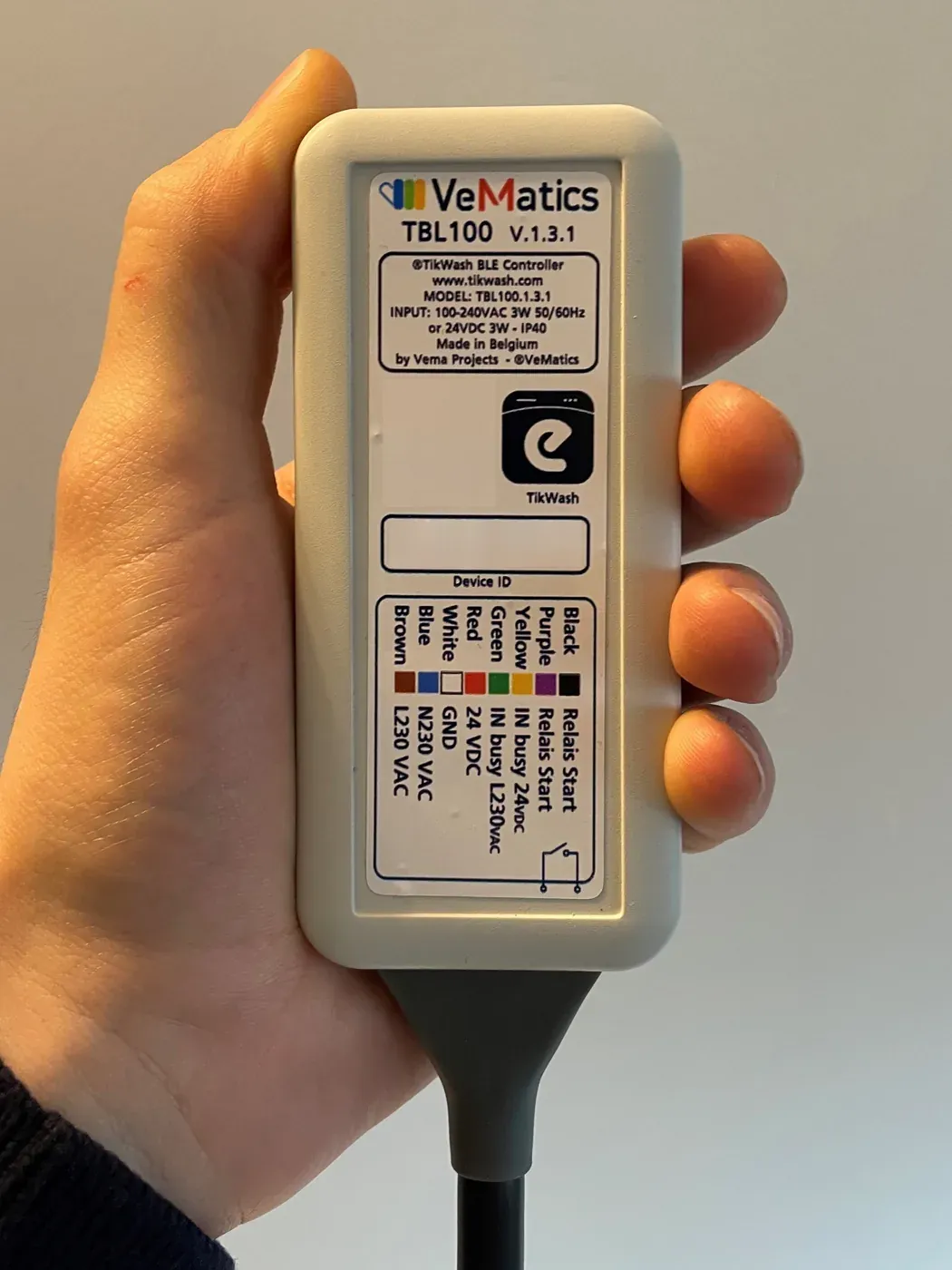 Een TBL100-module in de hand: een compacte lichtgrijze behuizing met het VeMatics- en TikWash-label, een aansluitschema en de kabel.